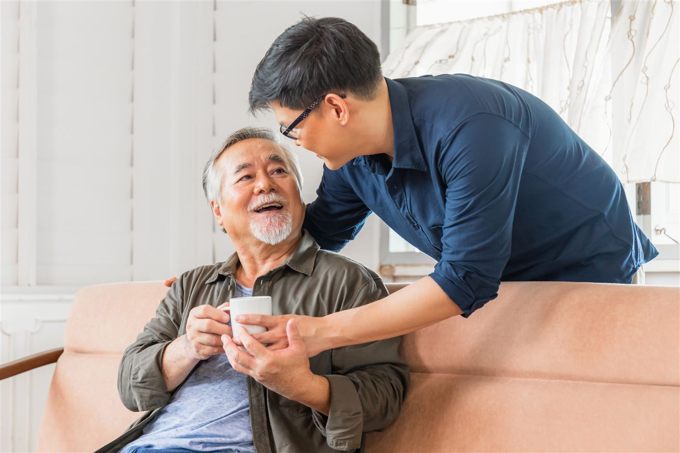 A caregiver leans in to hand a smiling senior man a cup of coffee on the sofa.
