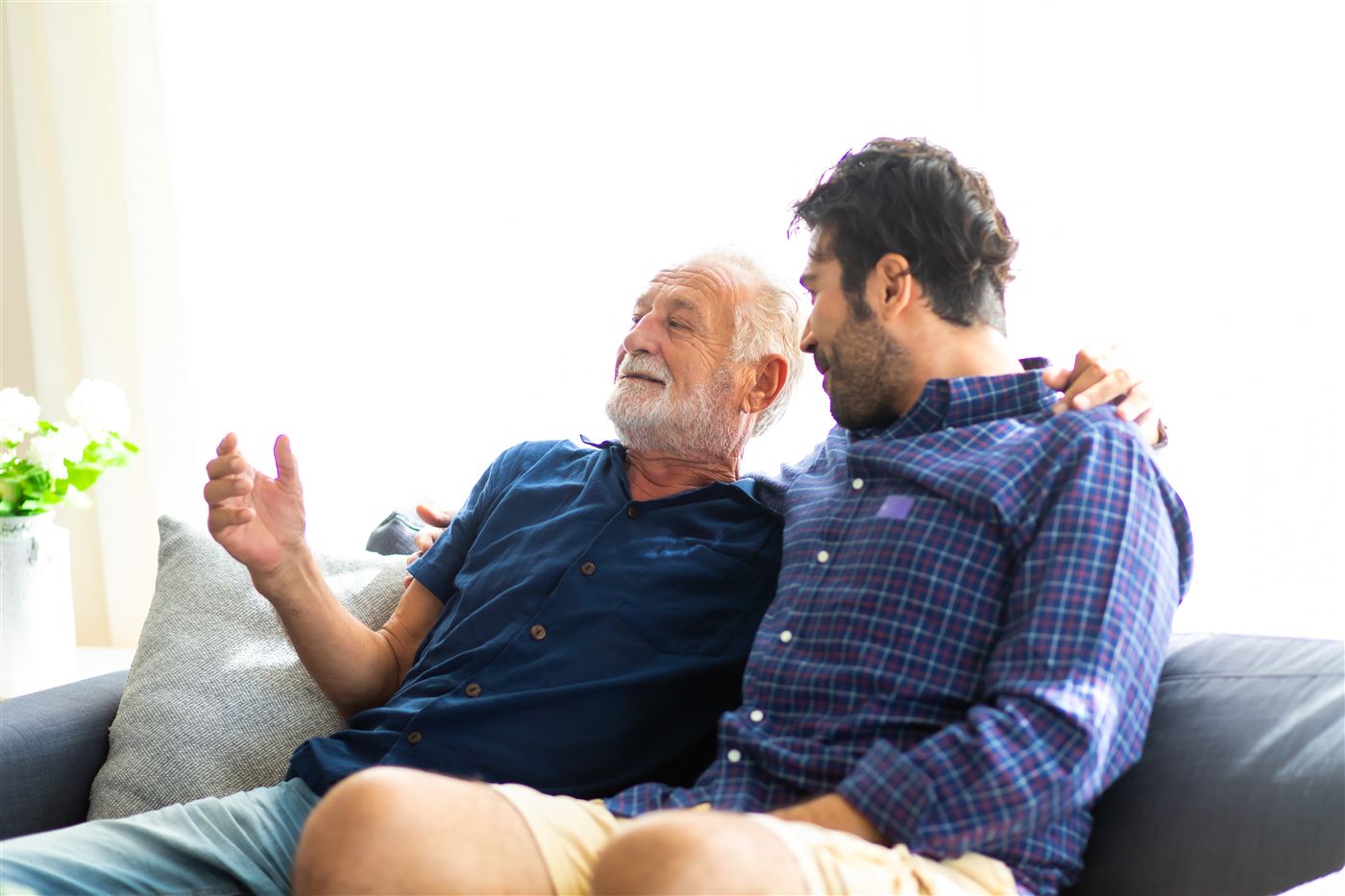A father and son sit close on a sofa at home, talking and smiling.