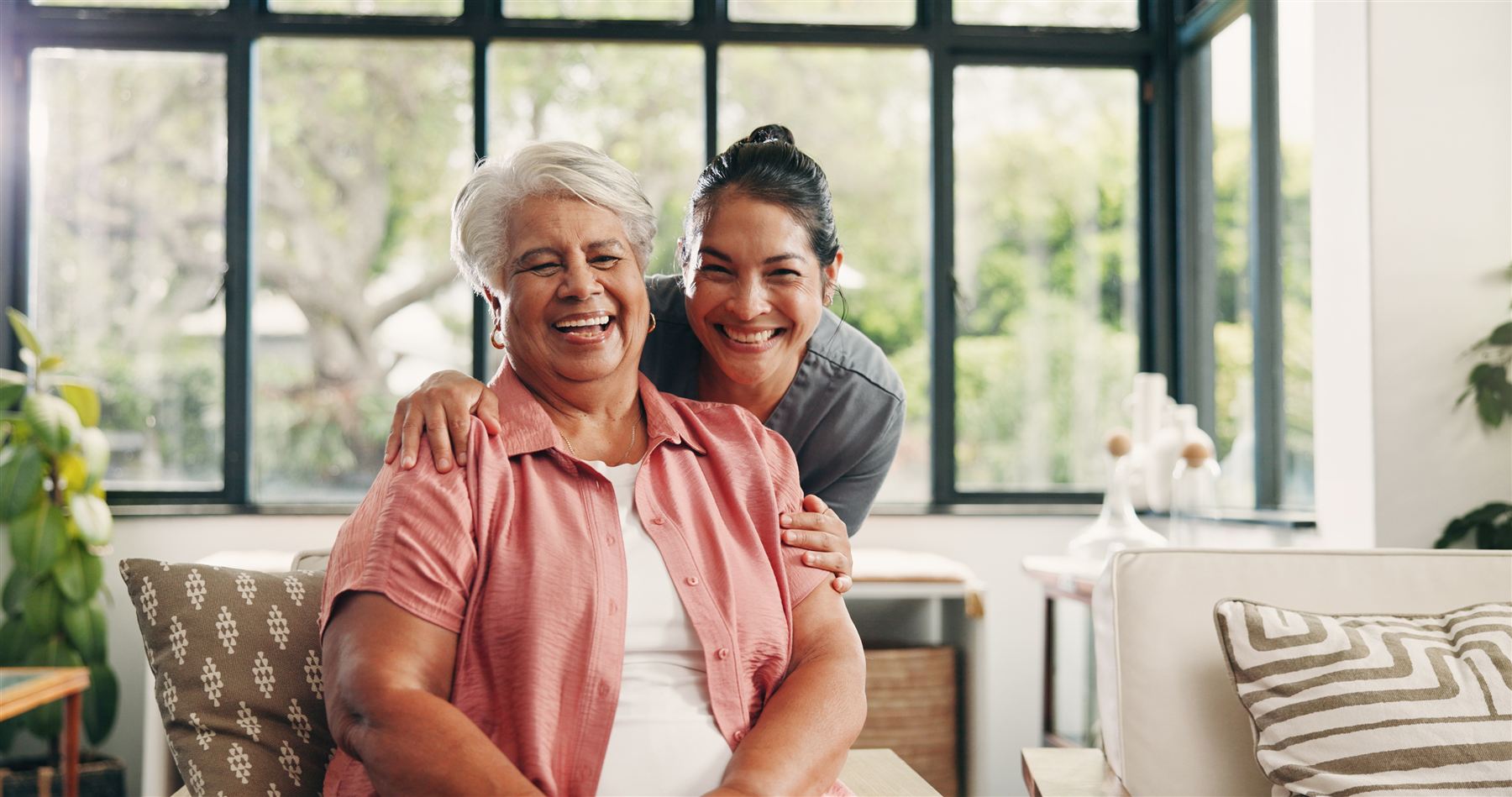 A senior woman laughing with her daughter, who has an arm around her shoulders, in a bright sunroom.