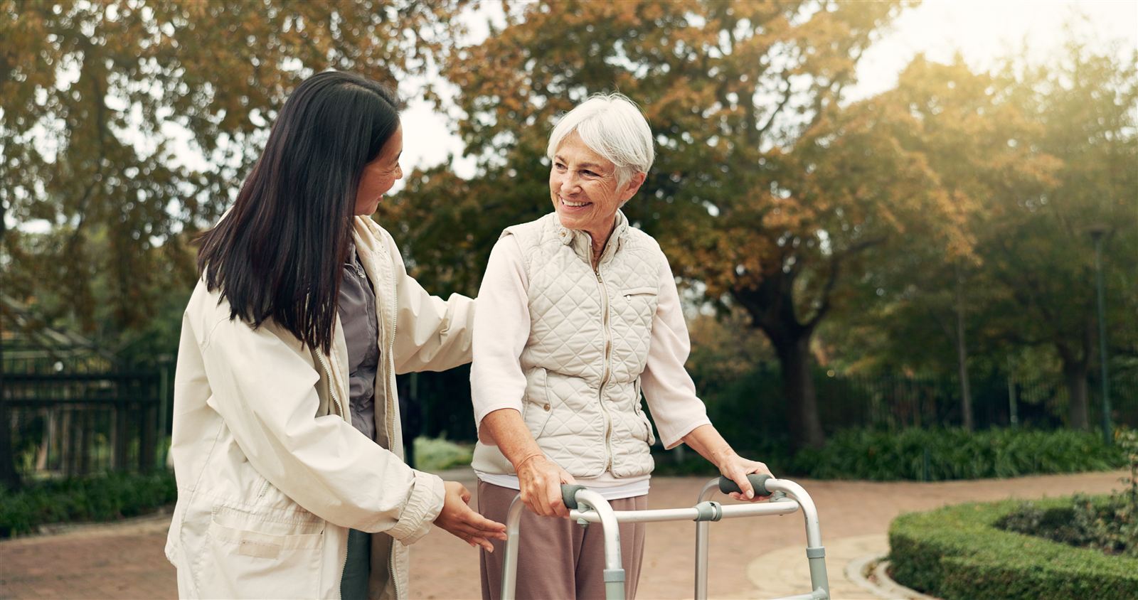 A caregiver walks beside a senior woman using a walker along a garden path in autumn.
